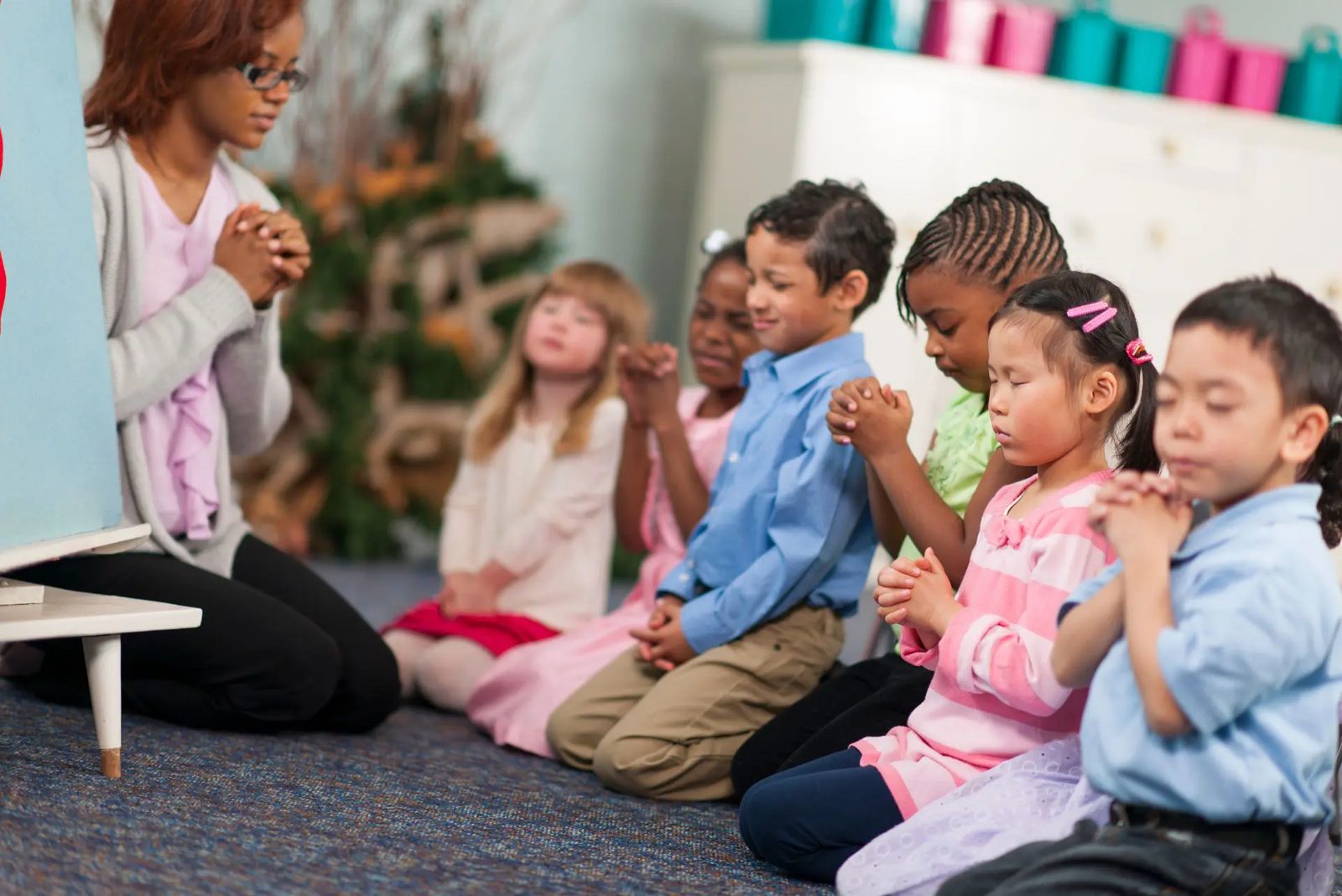 Children of many backgrounds kneeling and praying together with their Sunday School teacher