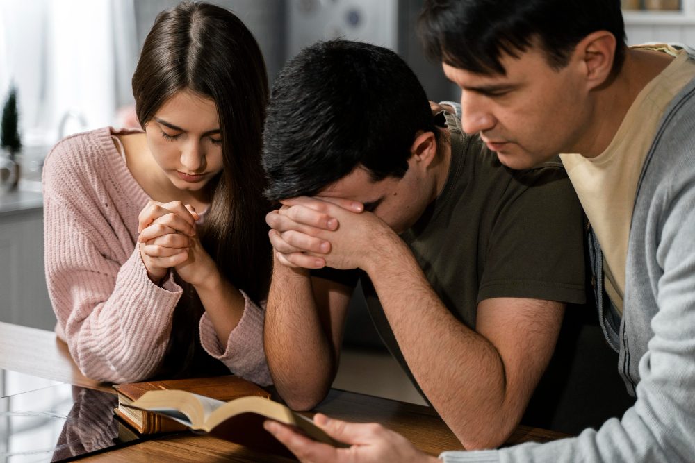 Three people praying together around an open Bible at a kitchen table