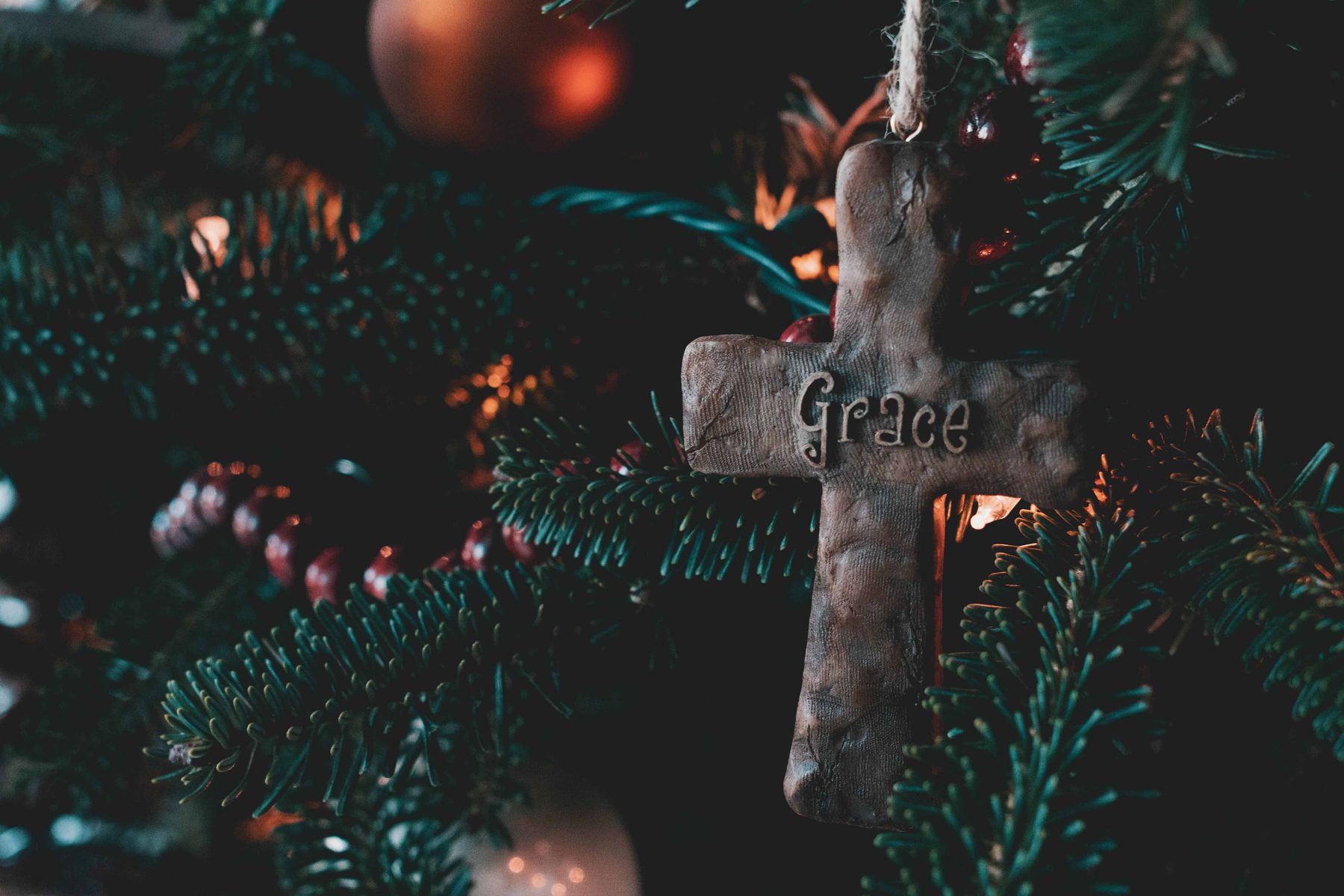 A wooden cross engraved with the word Grace, hanging among the branches and lights of a Christmas tree