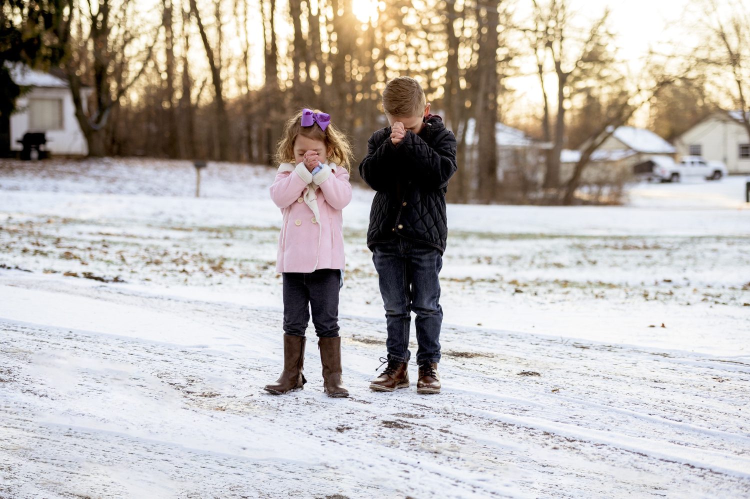 Two young children standing outdoors in the snow with their heads bowed in prayer at sunrise